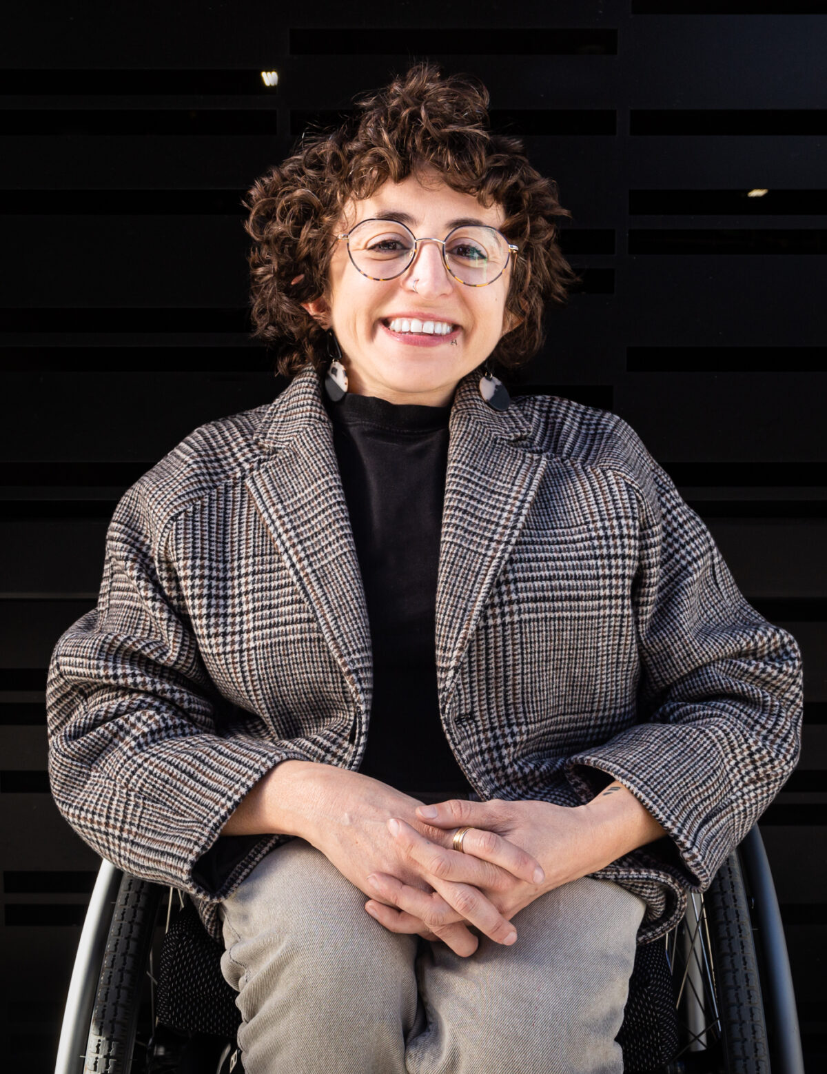 White queer person smling in their manual wheelchair with short brown hair, round glasses, earirings, and a blazer.