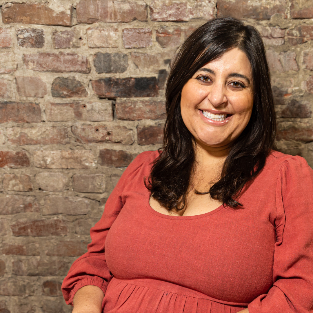 Annie is smiling in front of a brick background and wearing a terra cotta colored linen dress. She has medium length brown hair, brown eyes, and light olive skin.