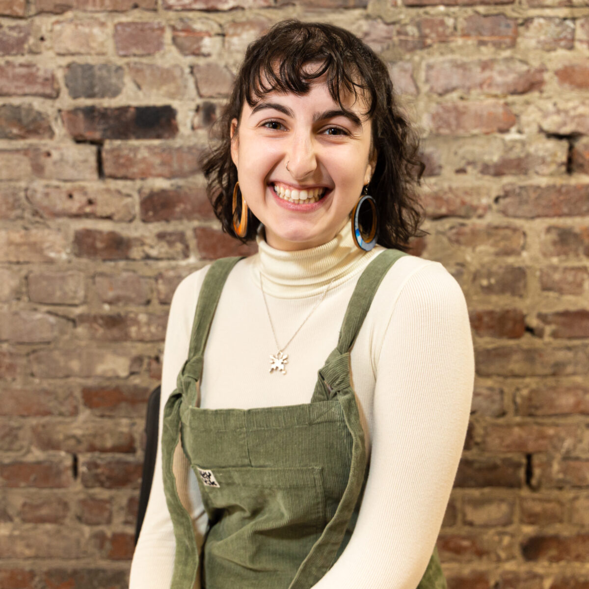 Ari is a white non-binary person with shoulder-length brown hair. They are sitting in front of a brick wall and smiling. They are wearing green overalls with a cream-colored turtleneck underneath, orange and blue hoop earrings, and a silver necklace with a butterfly pendant.