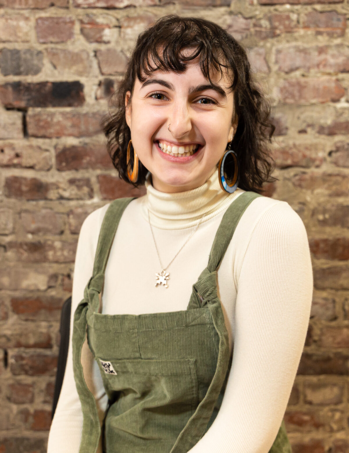 Ari is a white non-binary person with shoulder-length brown hair. They are sitting in front of a brick wall and smiling. They are wearing green overalls with a cream-colored turtleneck underneath, orange and blue hoop earrings, and a silver necklace with a butterfly pendant.