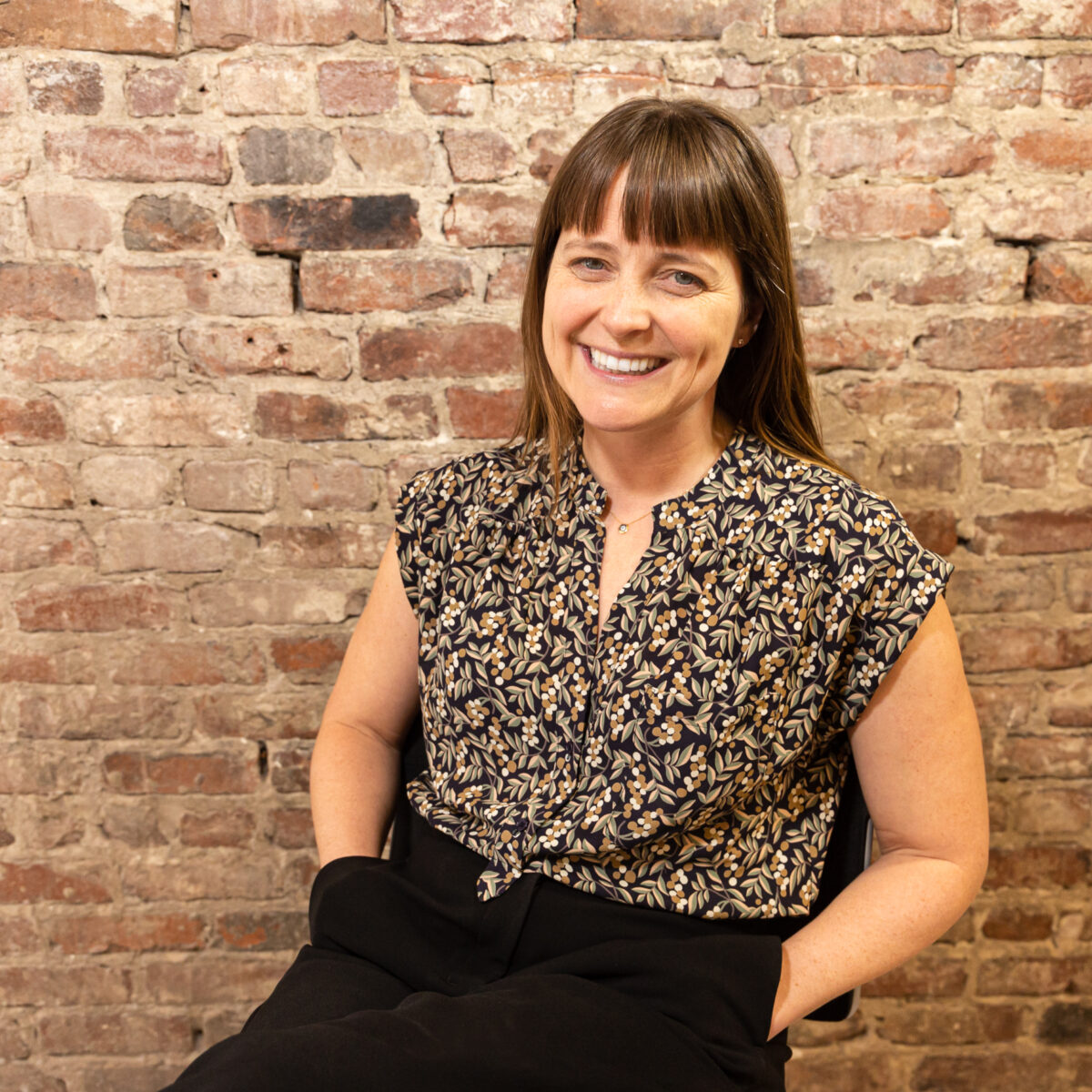 A white middle aged woman with brown hair is smiling and seated in front of a brick wall with her hands in her pockets.