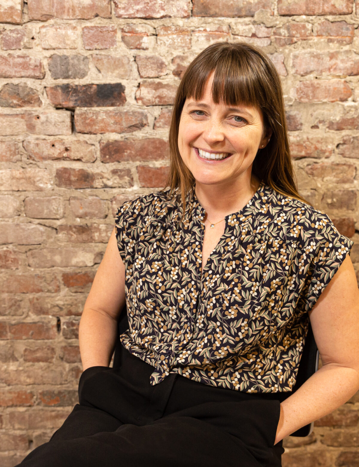 A white middle aged woman with brown hair is smiling and seated in front of a brick wall with her hands in her pockets.