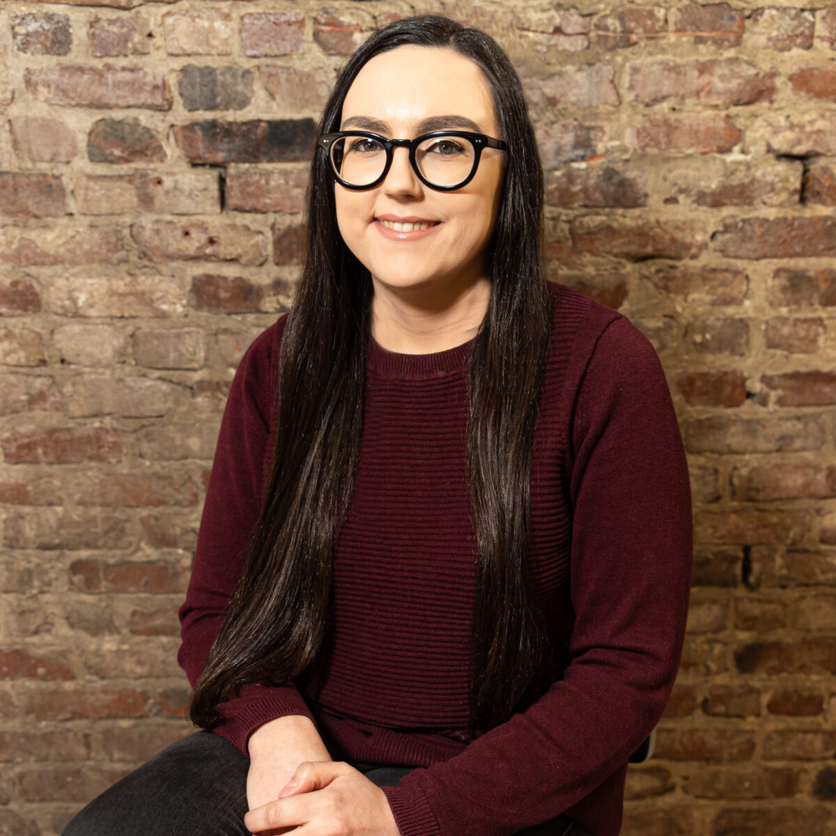 A light-skinned Native woman with long black hair, black glasses, and a burgundy shirt smiles in front of a brick background