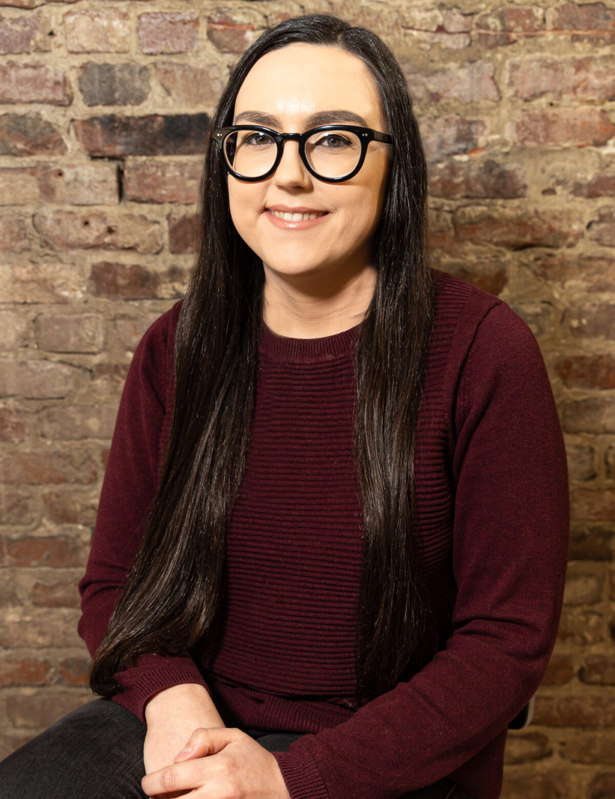 A light-skinned Native woman with long black hair, black glasses, and a burgundy shirt smiles in front of a brick background