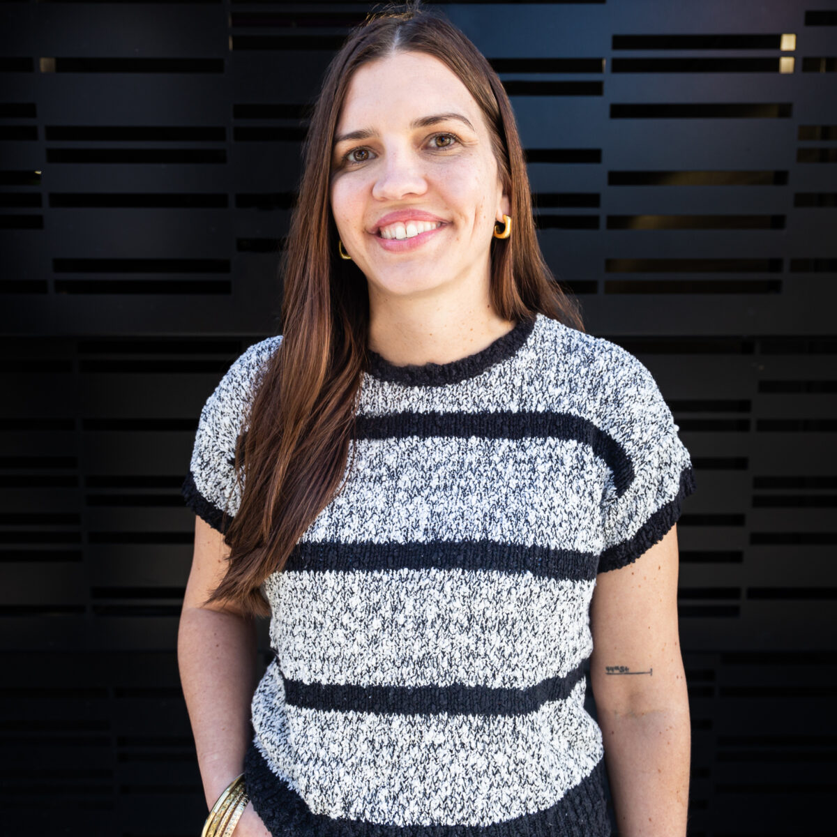 Katie is a white woman with long straight brown hair. Katie is standing in front of a black wall, smiling, wearing a black and gray striped shirt.