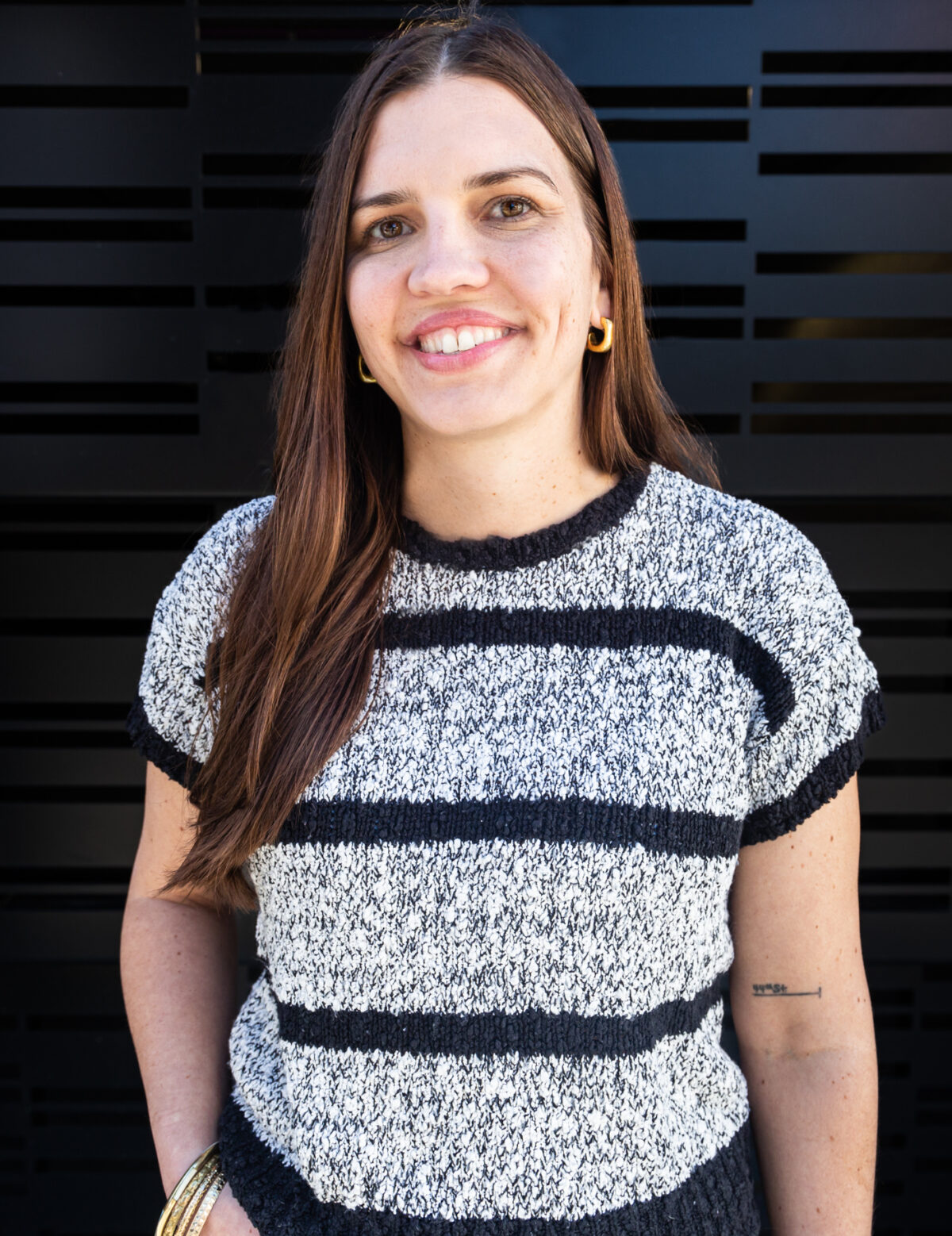 Katie is a white woman with long straight brown hair. Katie is standing in front of a black wall, smiling, wearing a black and gray striped shirt.