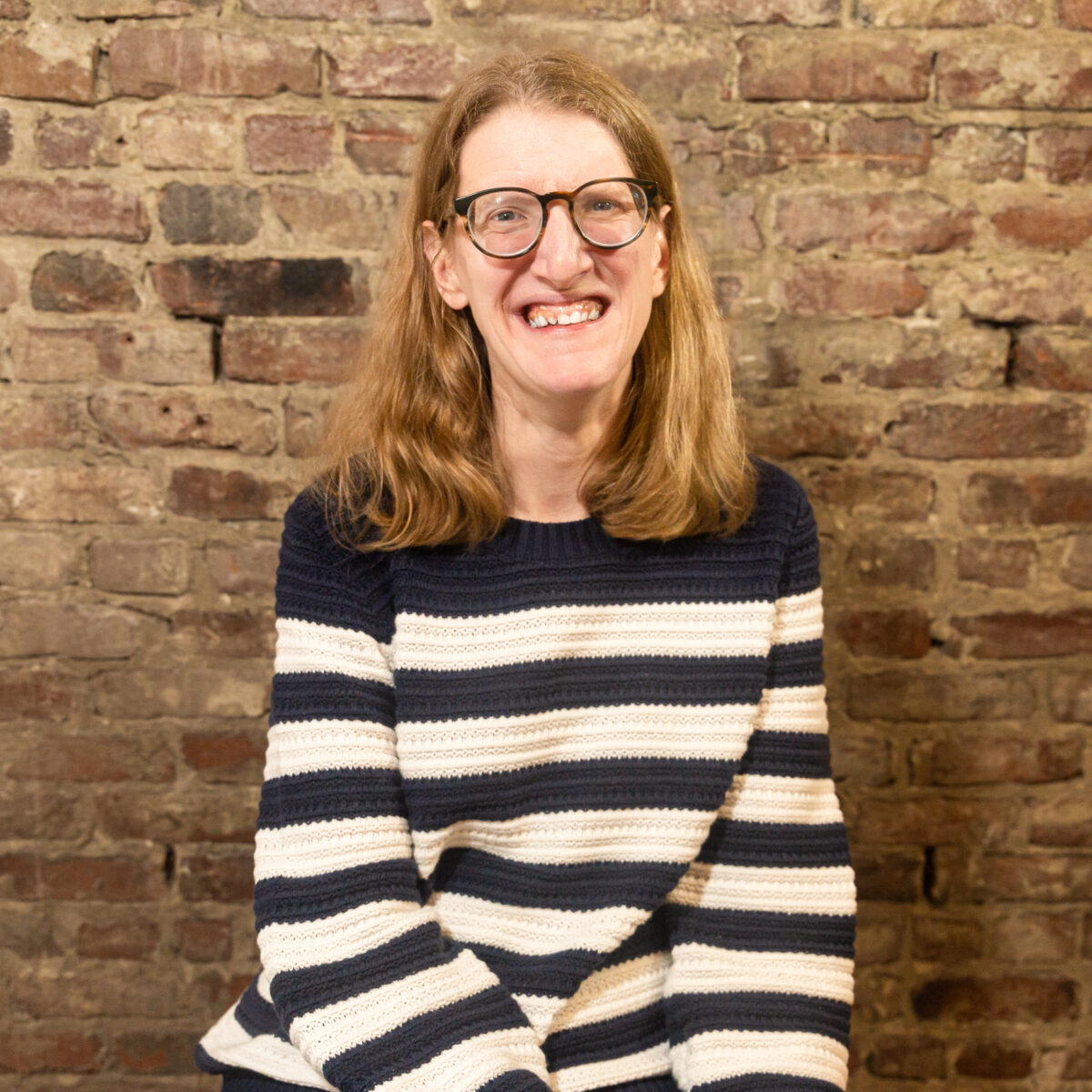 Molly, a white woman with blonde hair, is sitting in front of a brick wall. She is wearing glasses and a white and blue striped sweater.