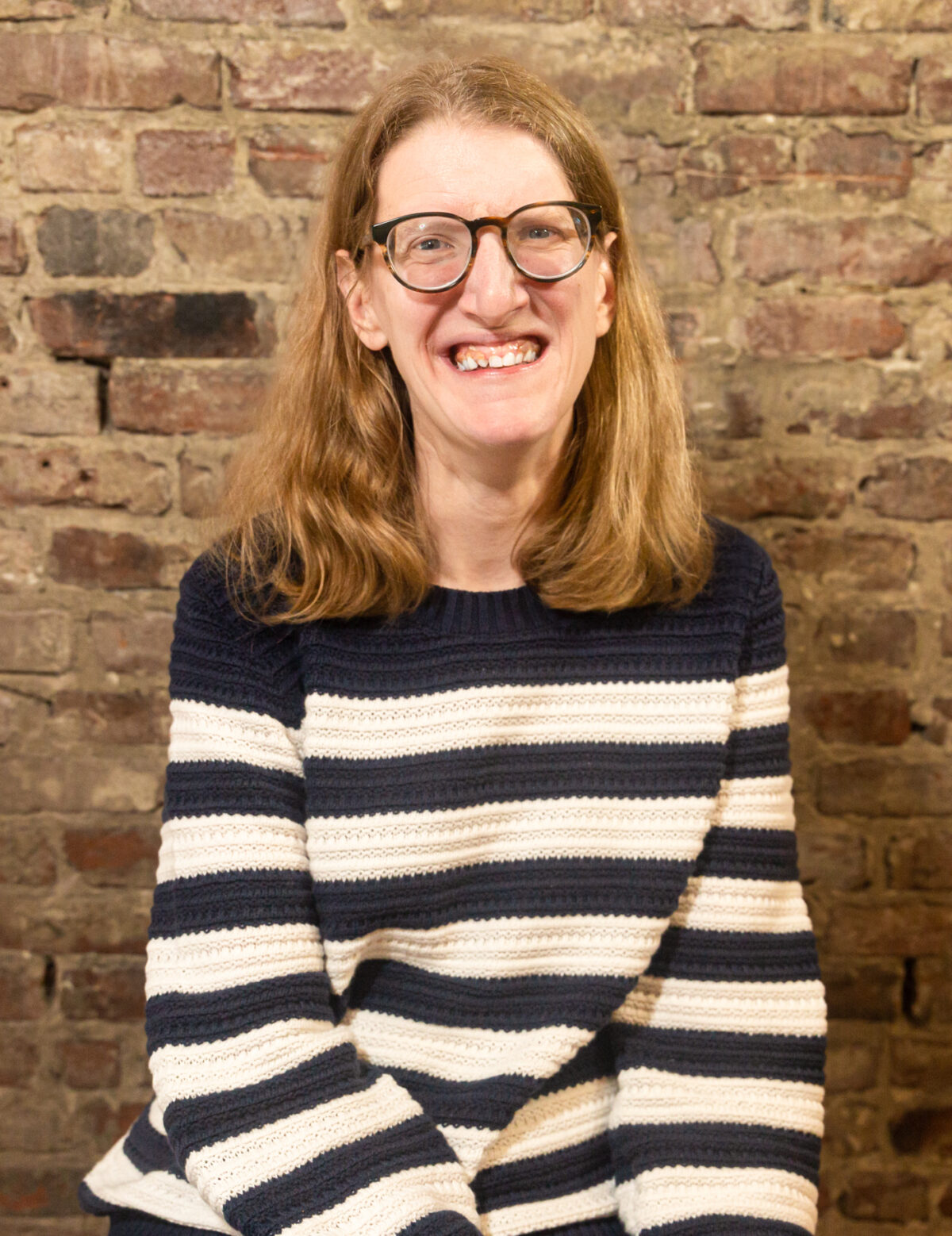 Molly, a white woman with blonde hair, is sitting in front of a brick wall. She is wearing glasses and a white and blue striped sweater.