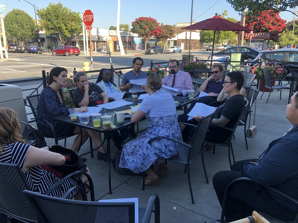 A table outside with people sitting around the table. There are papers, building design plans, on the table and being held by some of the people.