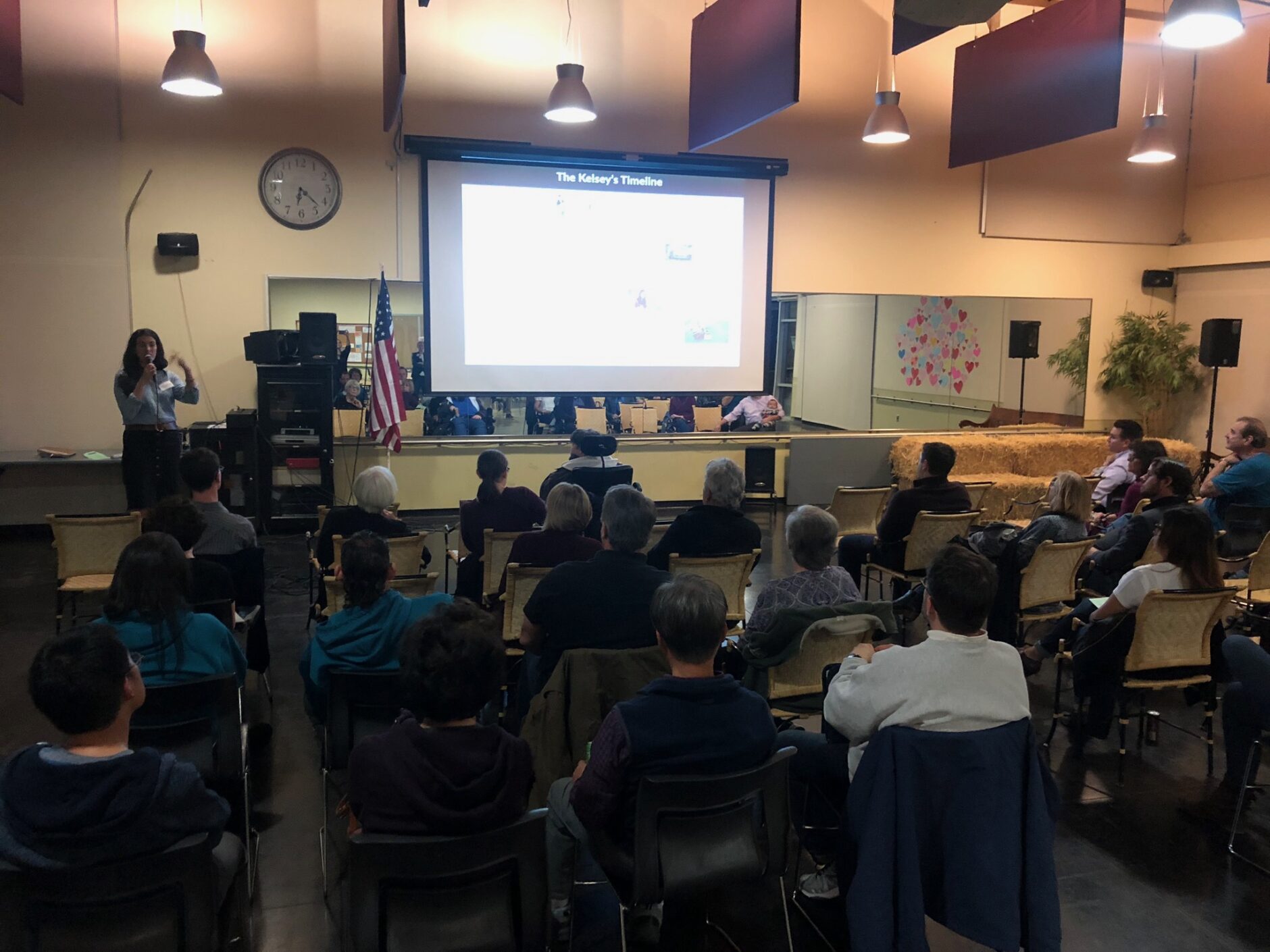 A community room with people seated in chairs and someone standing in front of the room with a slideshow screen showing project information.