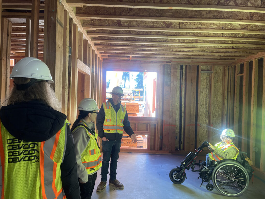 A group of 4 people in a construction site. They are wearing yellow vest and hard hats. Three are standing and one is using a wheelchair.