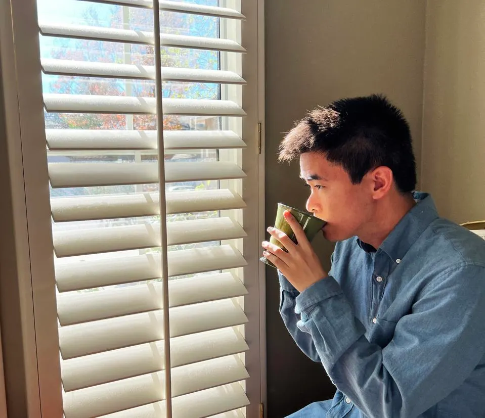 Justin Tsang, a young Asian man, is pictured holding a coffee cup looking through shutters. He has ark hair and is wearing a blue button down shirt.