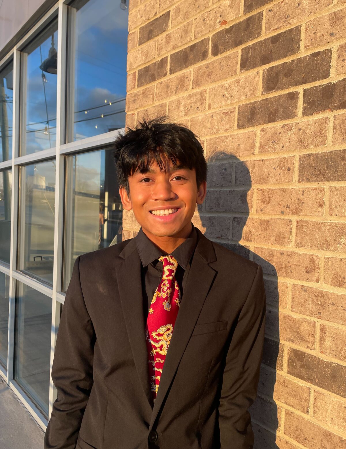 Brown-skin Southeast/South Asian male with shorter black hair and bangs, wearing a black blazer, a black button-up dress shirt underneath, and a red tie with gold Chinese dragons on it.