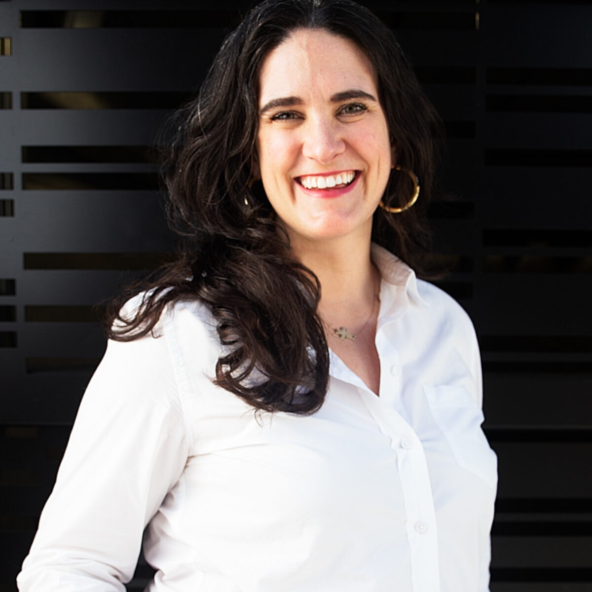 A white woman smiling at the camera with long, wavy, brown hair. She is wearing gold hoop earrings and a white button-up shirt standing in front of a black wall.