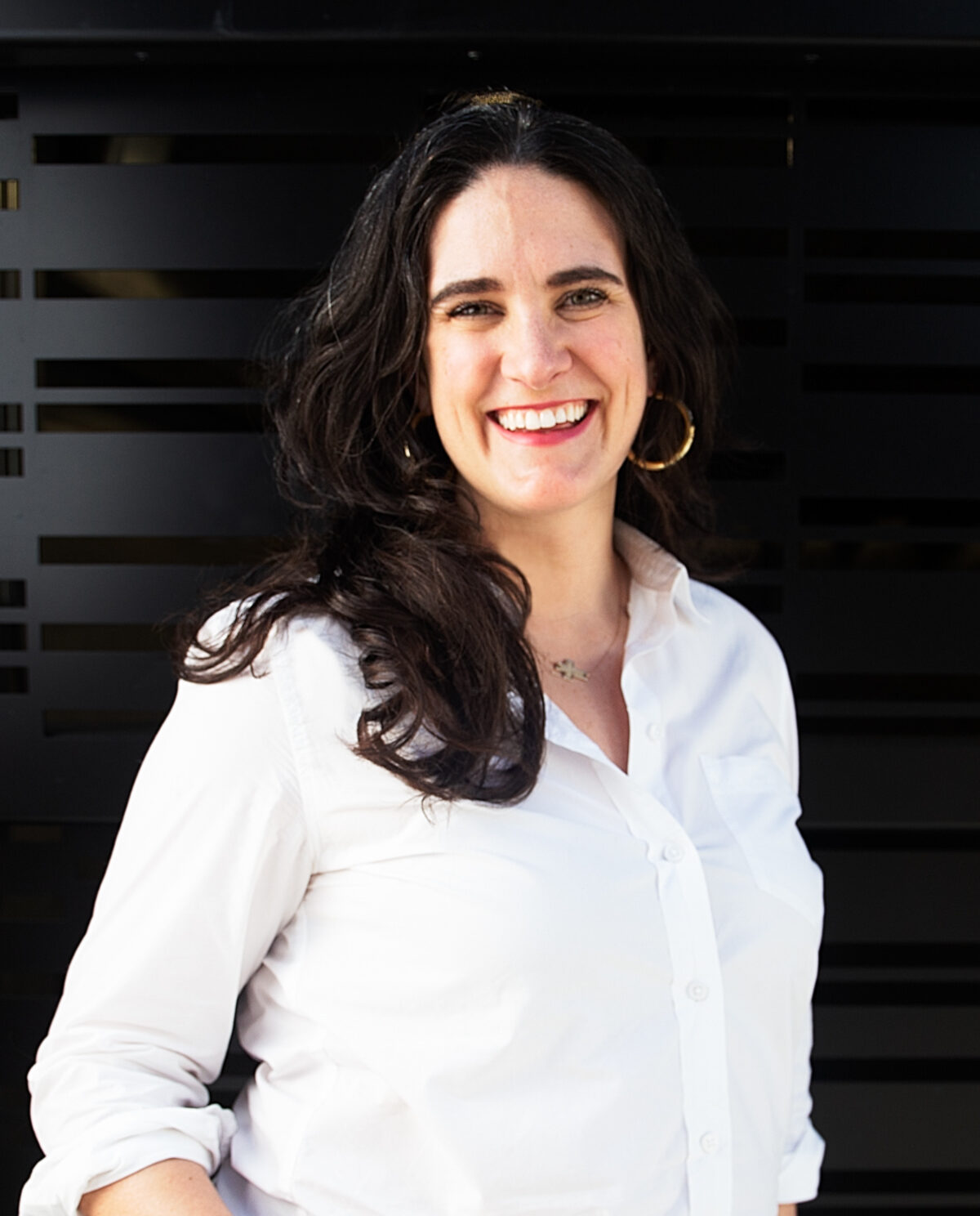 A white woman smiling at the camera with long, wavy, brown hair. She is wearing gold hoop earrings and a white button-up shirt standing in front of a black wall.