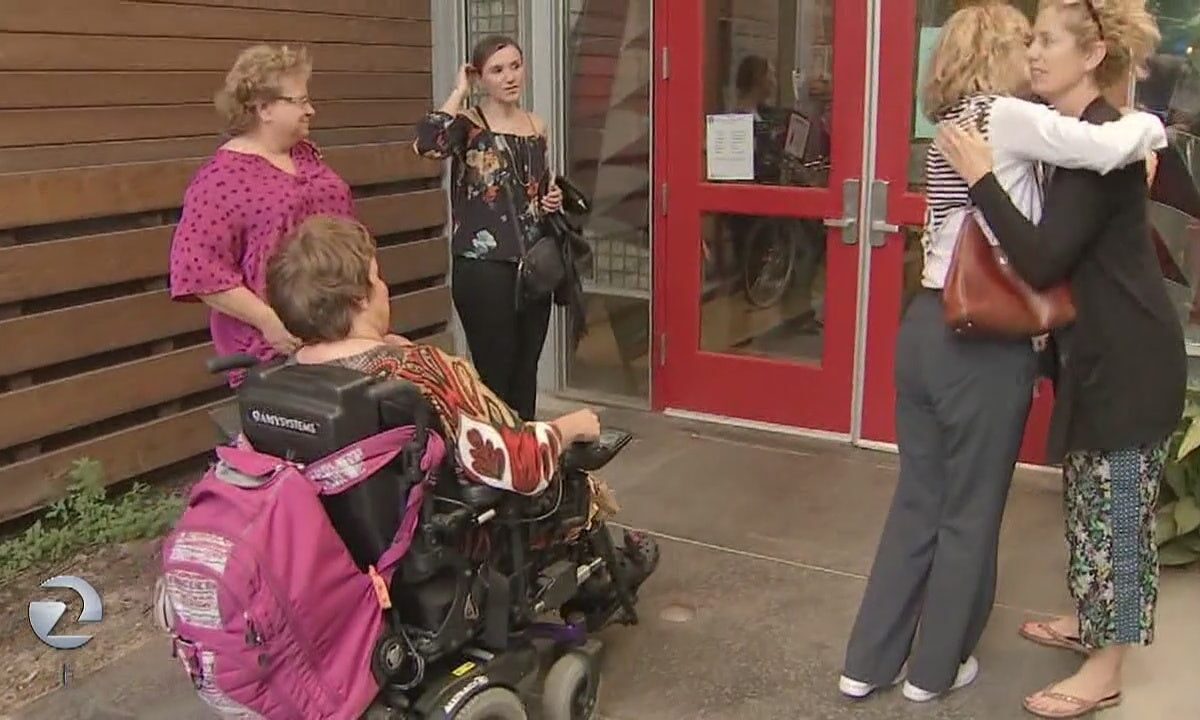 Two people outside of a doorway hug, as three other people, one who is in a wheelchair, look on.