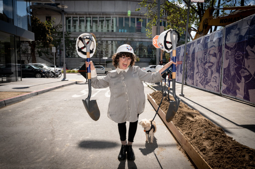 Allie Cannington, a white person with short, brown, curly hair wearing a hard hat, a striped shirt and black pants stands smiling enthusiastically holding construction shovels in each of their hands. 