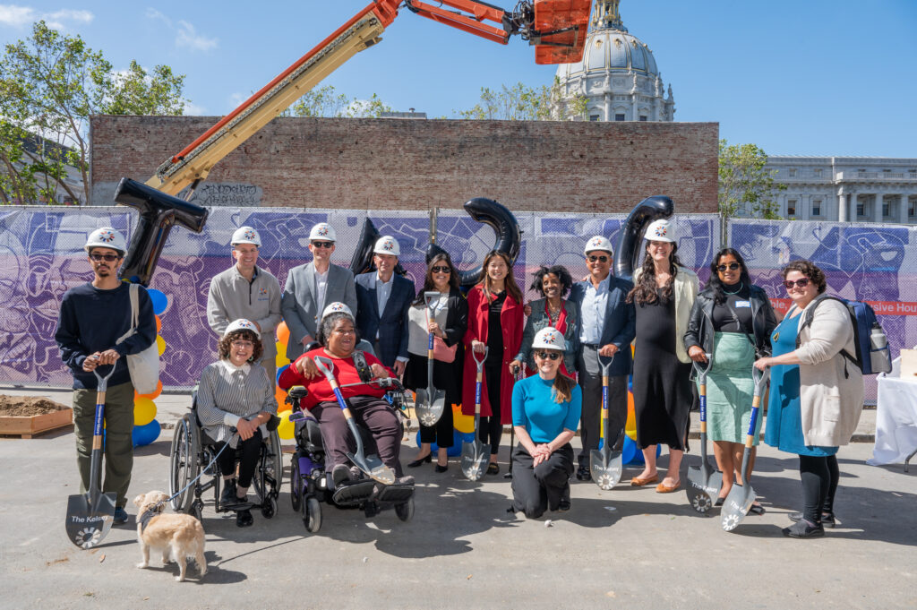 A group of people, some holding construction shovels, sit and stand against the wall of the construction site of The Kelsey Civic Center. A construction crane and the dome of the San Francisco Civic Center is visible in the background.