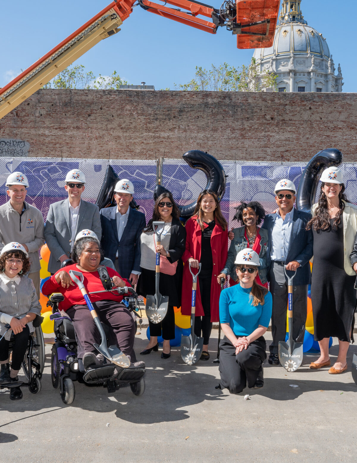 A group of people with and without disabilities, some holding construction shovels and others wearing hard hats, sit and stand against the wall of the construction site of The Kelsey Civic Center. A construction crane and the dome of the San Francisco Civic Center is visible in the background.