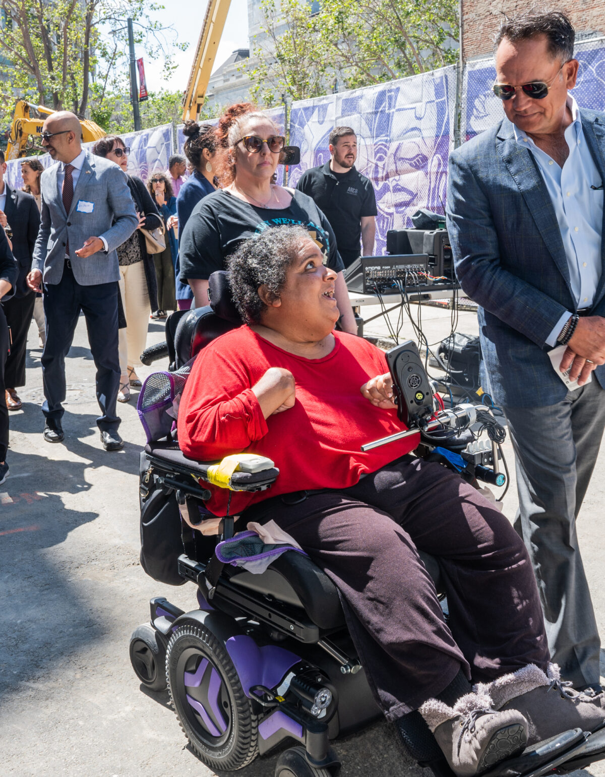 A white man and a black woman, one walking and one wheeling, in front of a crowd at The Kelsey Civic Center groundbreaking. There is a construction fence in the background.