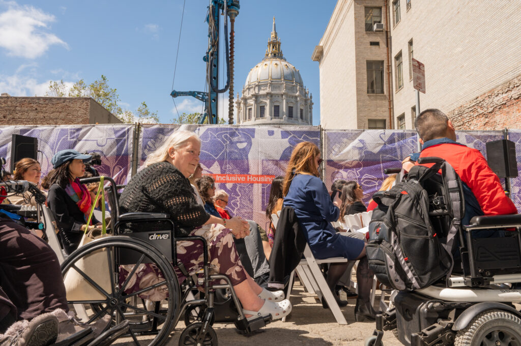 People sitting in chairs and wheelchairs at the groundbreaking ceremony.  The Civic Center dome and the wall of The Kelsey Civic Center construction site are visible in the background.