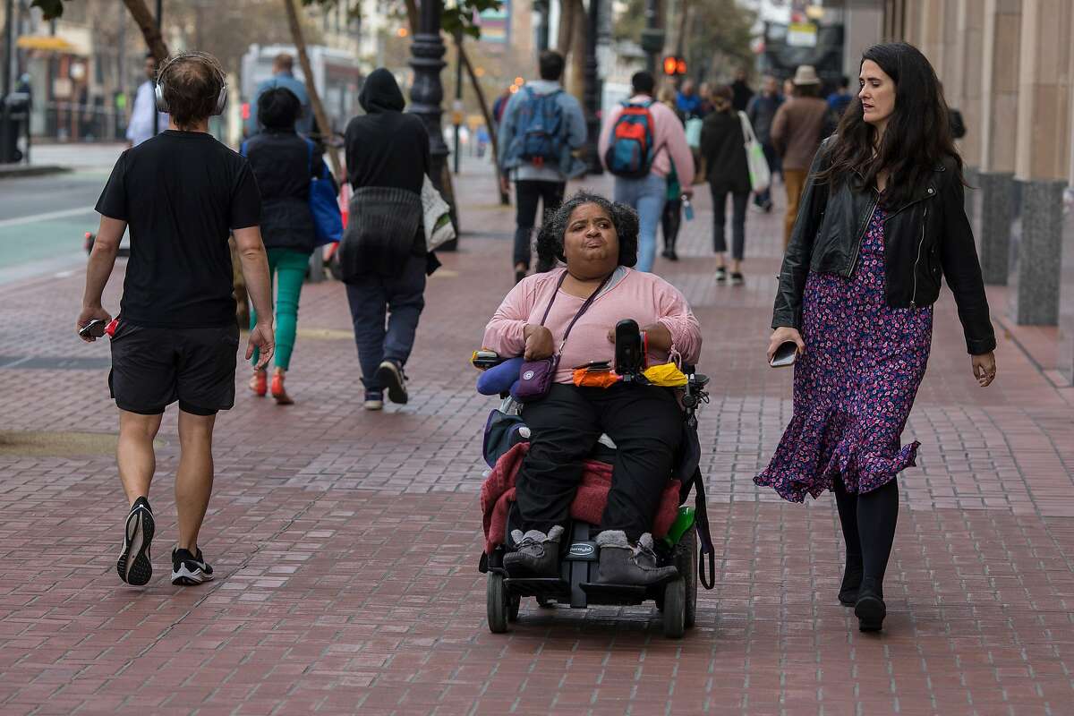 Micaela Connery a light skinned woman with long dark hair wearing a purple dress is seen walking with Liz Grigsby, a dark skinned woman in a wheelchair wearing a pink sweater and dark pants.