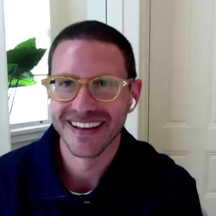 A light skinned person with a beard wearing glasses and a blue shirt. A plant is visible behind them.