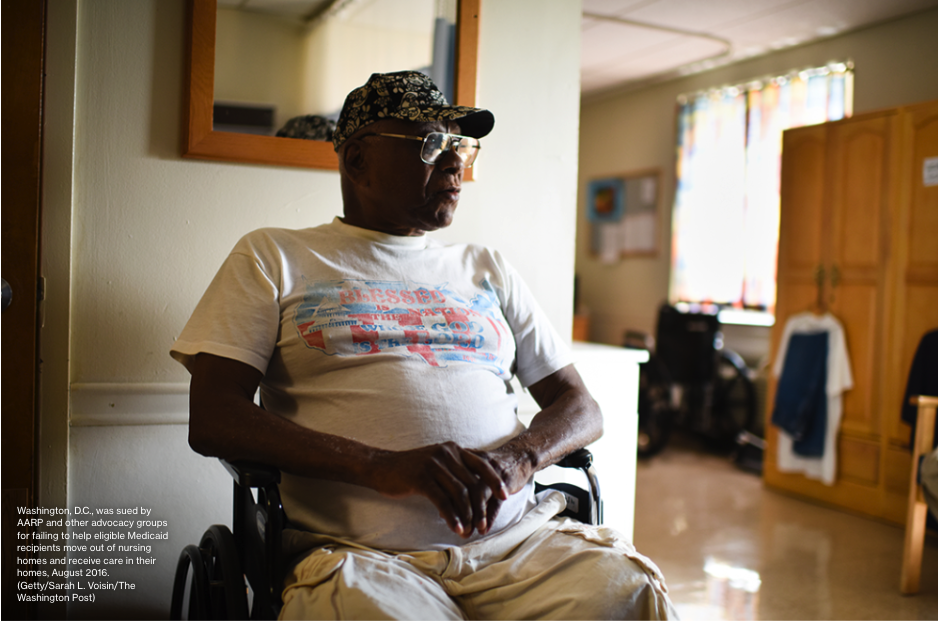 An Image of a dark-skinned man sitting in a wheelchair. He is wearing a hat, glasses, a t-shirt and light-colored pants. In the background a window and clothes hanging on a cabinet can be seen.