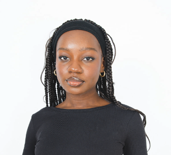 A dark-skinned Black woman with long, dark brown box braids is softly smiling in front of a white background. She is wearing a black, cloth headband, small gold hoop earrings, and a long sleeve black shirt.