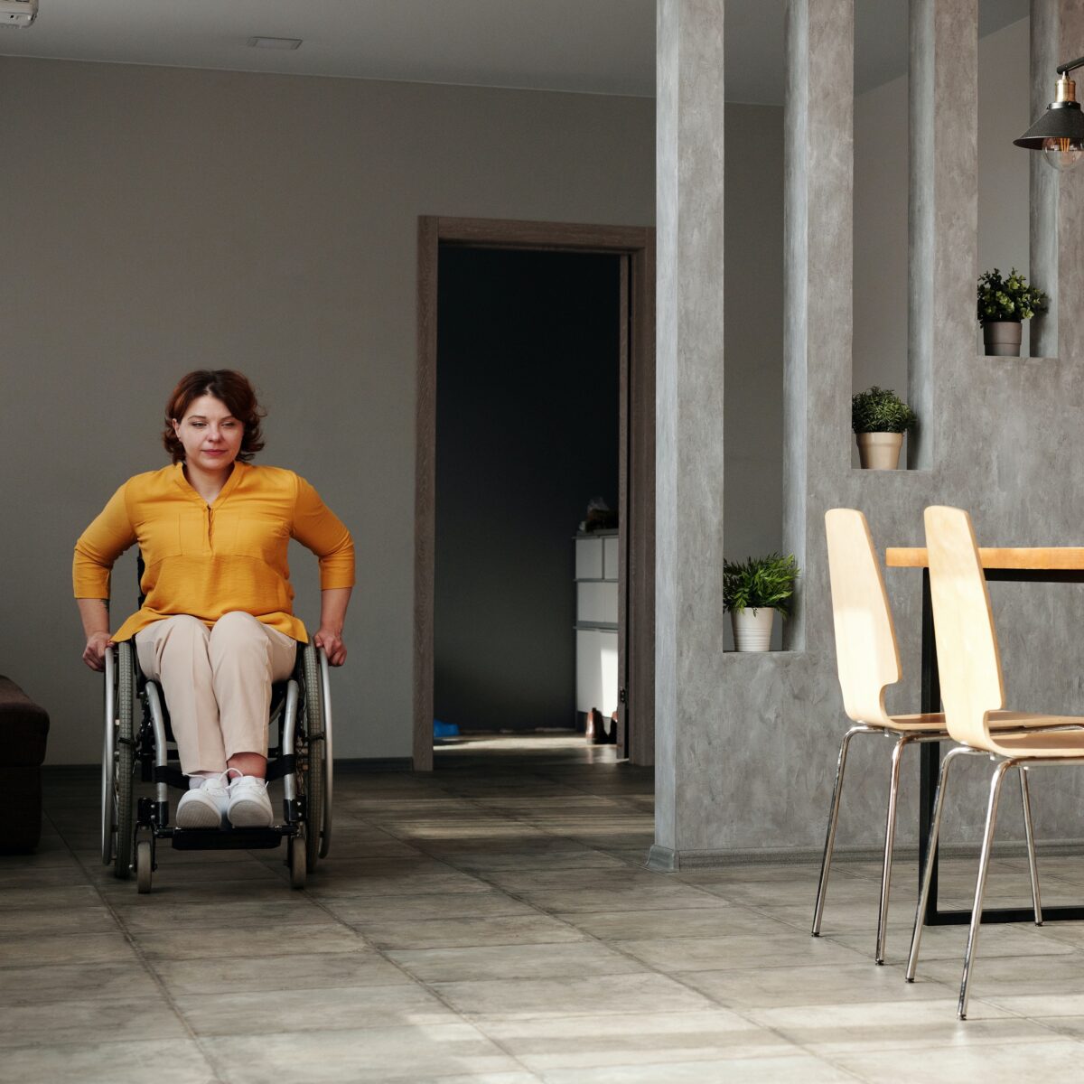 A light skinned woman wearing an orange button down shirt and tan pants wheels through a room in a wheelchair.