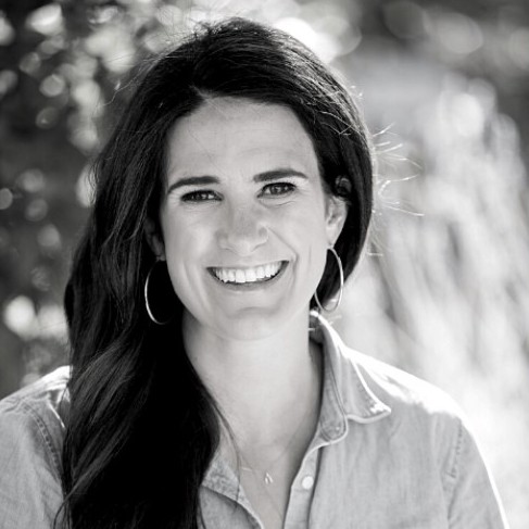 A black and white photo of Micaela Connery, a light skinned woman with long dark hair. She is smiling and wearing earings and a button-down shirt. with a collar