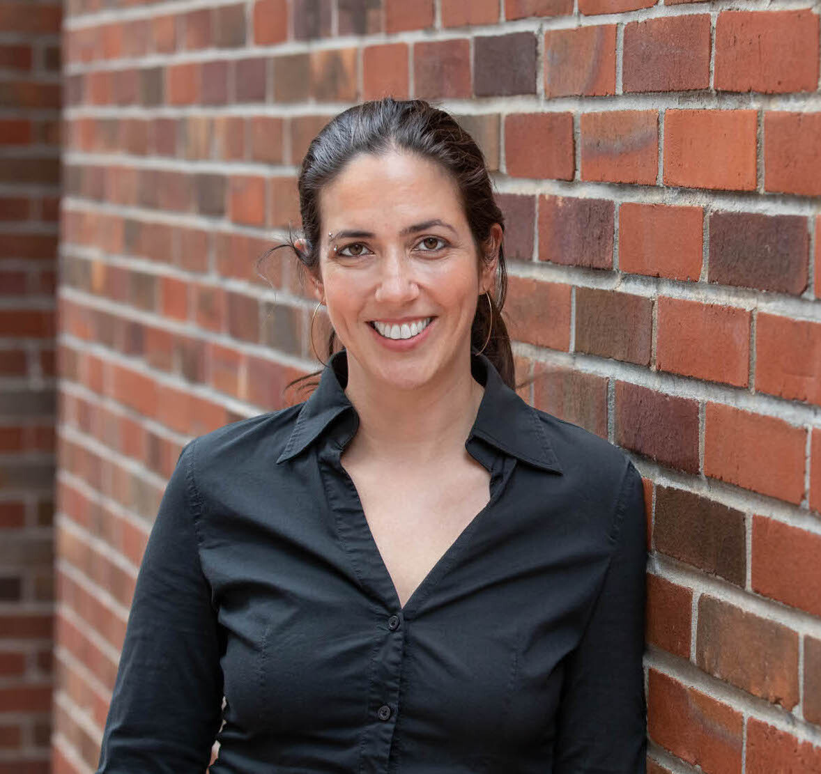 Louisa, a white woman with dark hair in a ponytail is smiling and standing in front of a red brick wall. She is wearing a black button-up shirt and gold earrings.