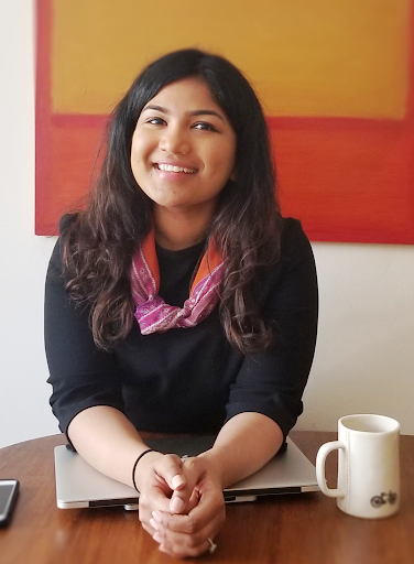 Arpitha Shetty, a dark-skinned woman with long dark hair, sits at a table. She is wearing a black shirt and a pink scarf. Her arms rest on a closed laptop computer. A painting is visible on the wall in the background behind her.