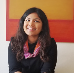 Arpitha is a dark-skinned Indian woman. She is wearing a black shirt and a pink sarf.