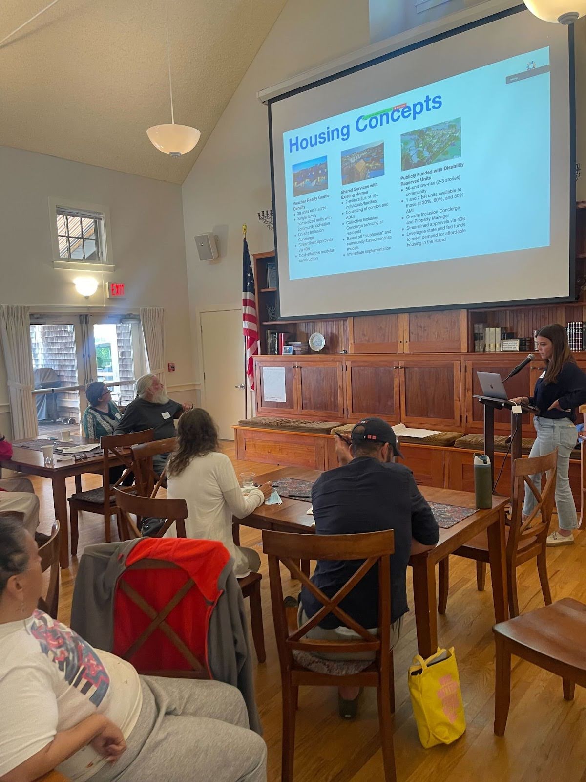 People sit in chairs and watch as Katie McAuley, Housing Development Manager at The Kelsey, gives a presentation. A slide about housing concepts is projected onto a screen.