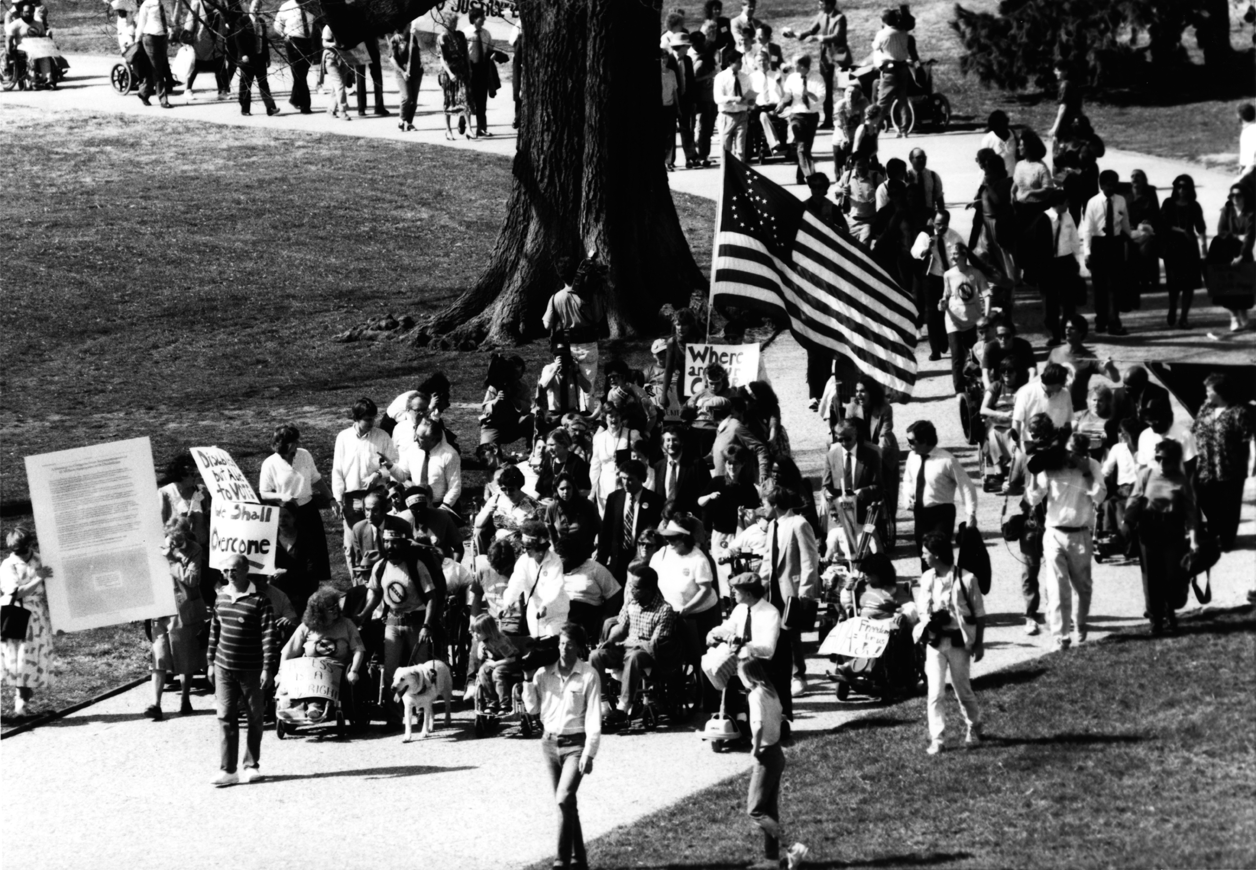 A black and white image of people with and without disabilities marching on a paved path near a tree and grass. One person holds an American flag with the stars in the shape of a wheelchair.