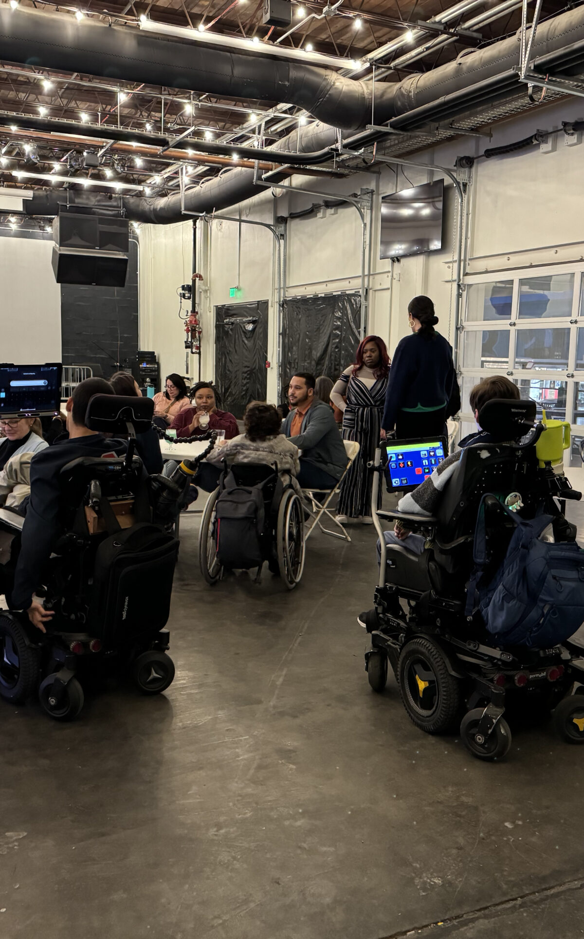 People sit in chairs and wheelchairs at tables in a large room with light colored walls.