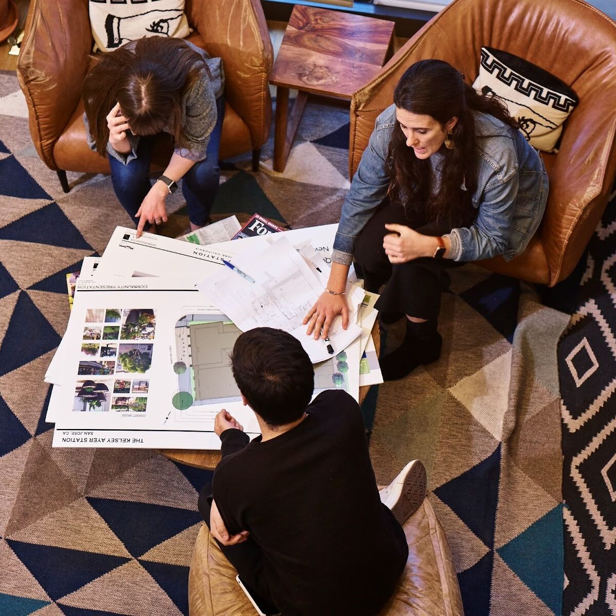 Three people are pictured sitting in armchairs, talking around a coffee table with floor plans on it.