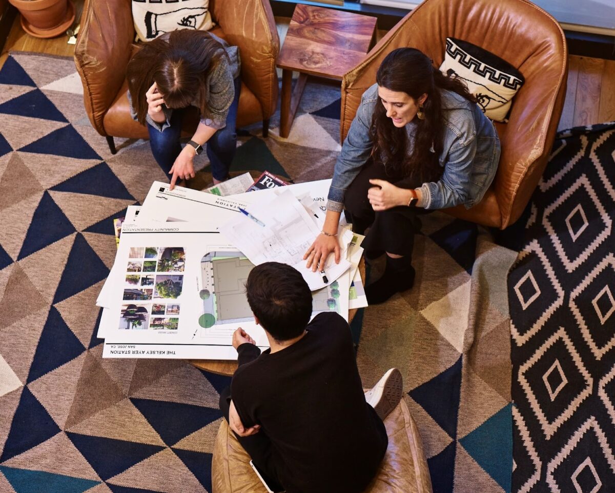 Three people are pictured sitting in armchairs, talking around a coffee table with floor plans on it.