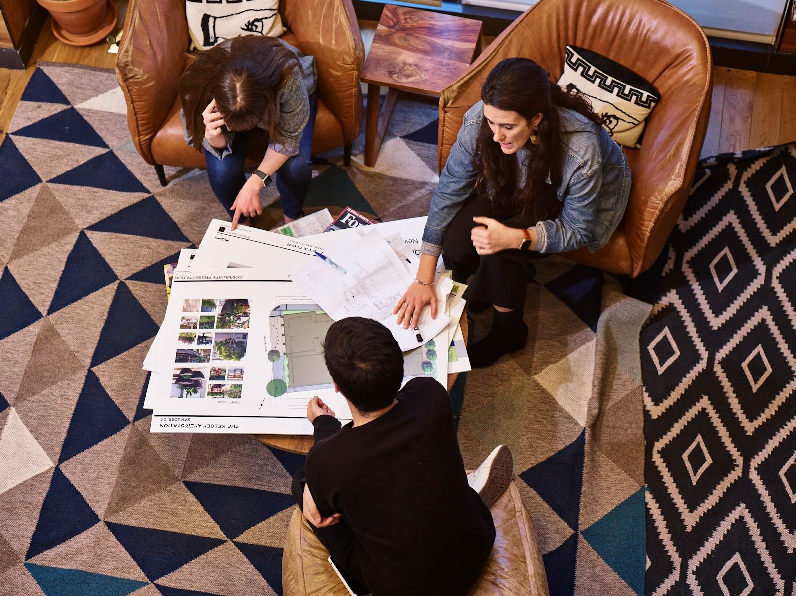 Three people are pictured sitting in armchairs, talking around a coffee table with floor plans on it.