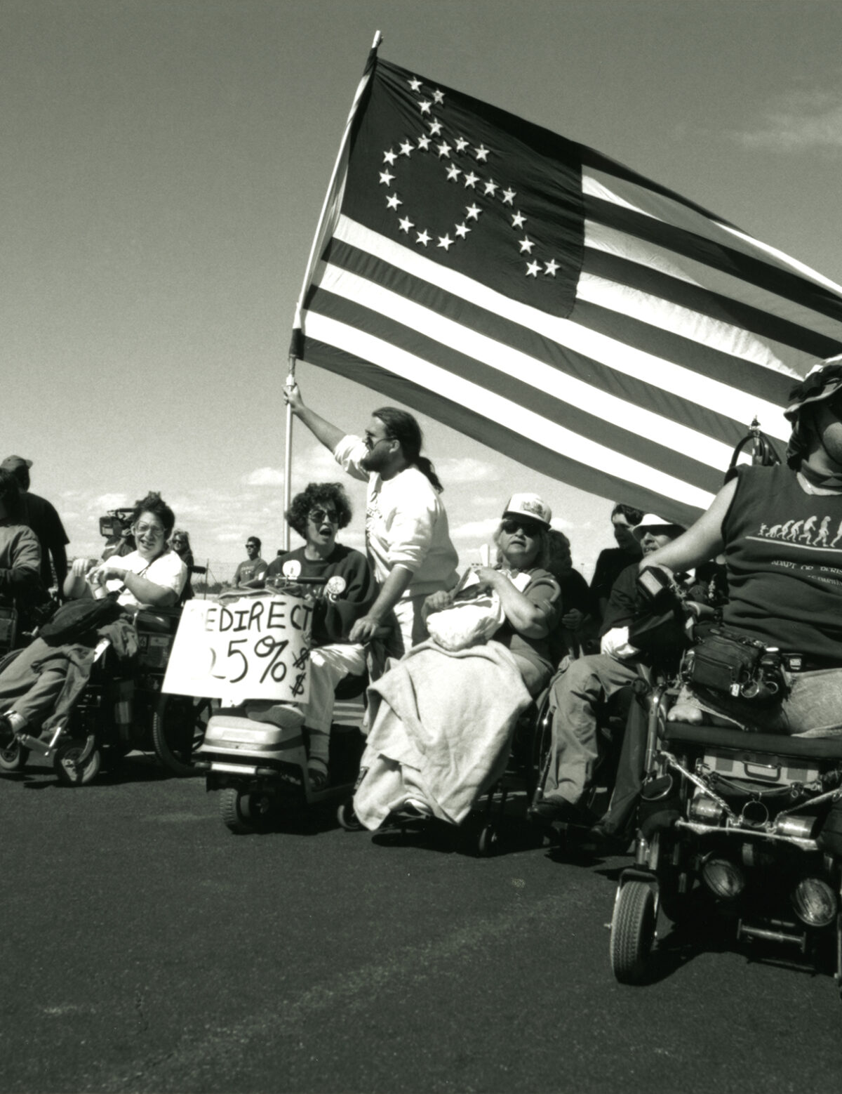 People sit in a line of wheelchairs. A person is seen holding an American Flag with the wheelchair symbol on it made out of stars