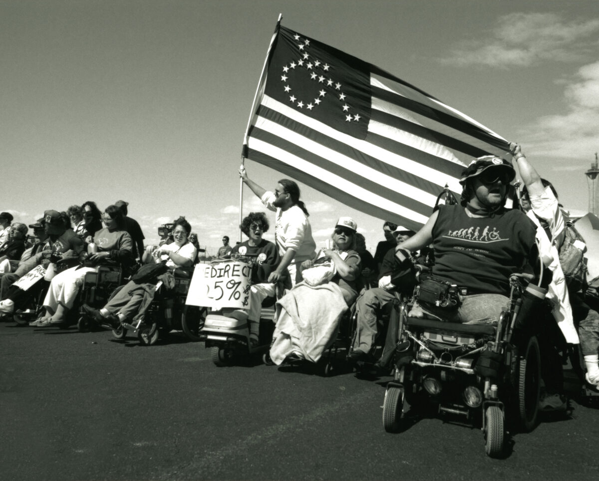 People sit in a line of wheelchairs. A person is seen holding an American Flag with the wheelchair symbol on it made out of stars