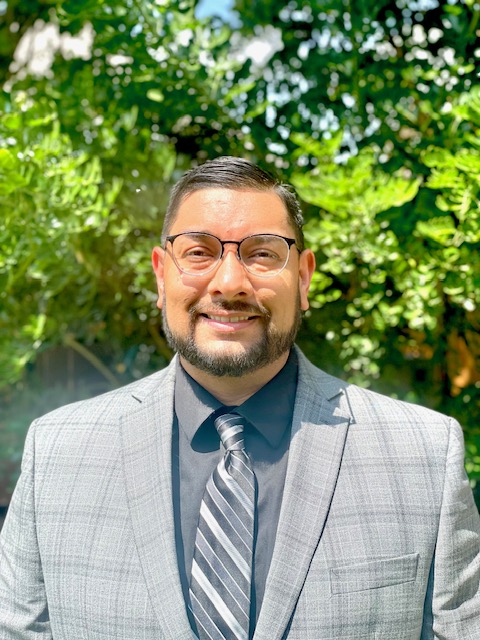 Headshot of a hispanic man with glasses in a gray suit with a black/gray tie smiling in front of a texas mountain laurel tree.