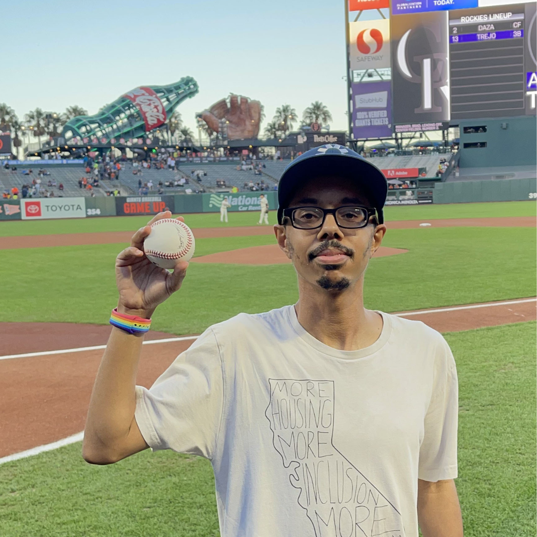 Isaac Haney-Owens is pictured on a baseball field holding a baseball. He is a dark skinned man with a beard and glasses.