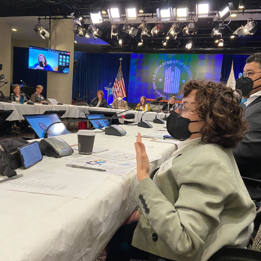 People with and without face masks sit in a circle at tables with microphones at the U.S. Department of Housing and Urban Development. The person closest to the camera is speaking into their microphone. They have short, curly hair and are wearing a tan blazer.