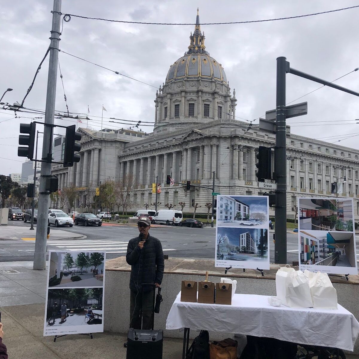 Isaac Haney Owens speaks in front of San Francisco City Hall. There is a table on one side of him and renderings of The Kelsey Civic Center behind him