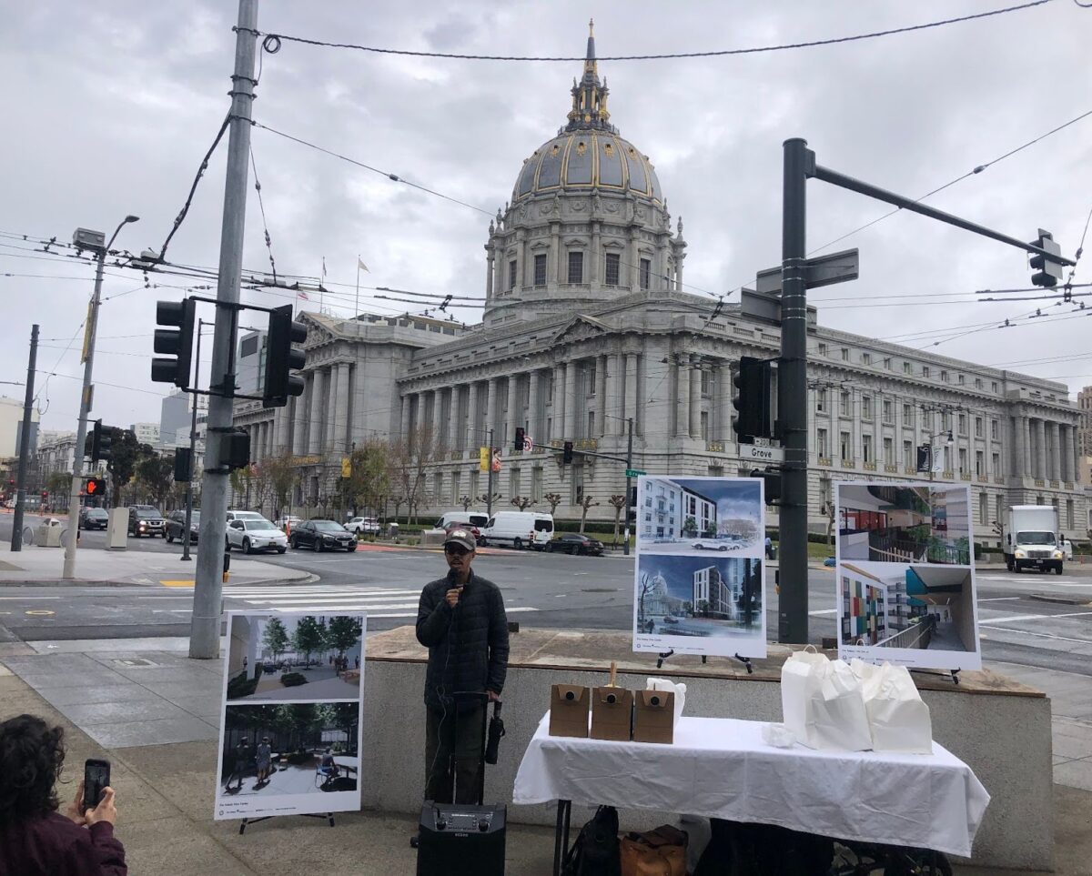 Isaac Haney Owens speaks in front of San Francisco City Hall. There is a table on one side of him and renderings of The Kelsey Civic Center behind him