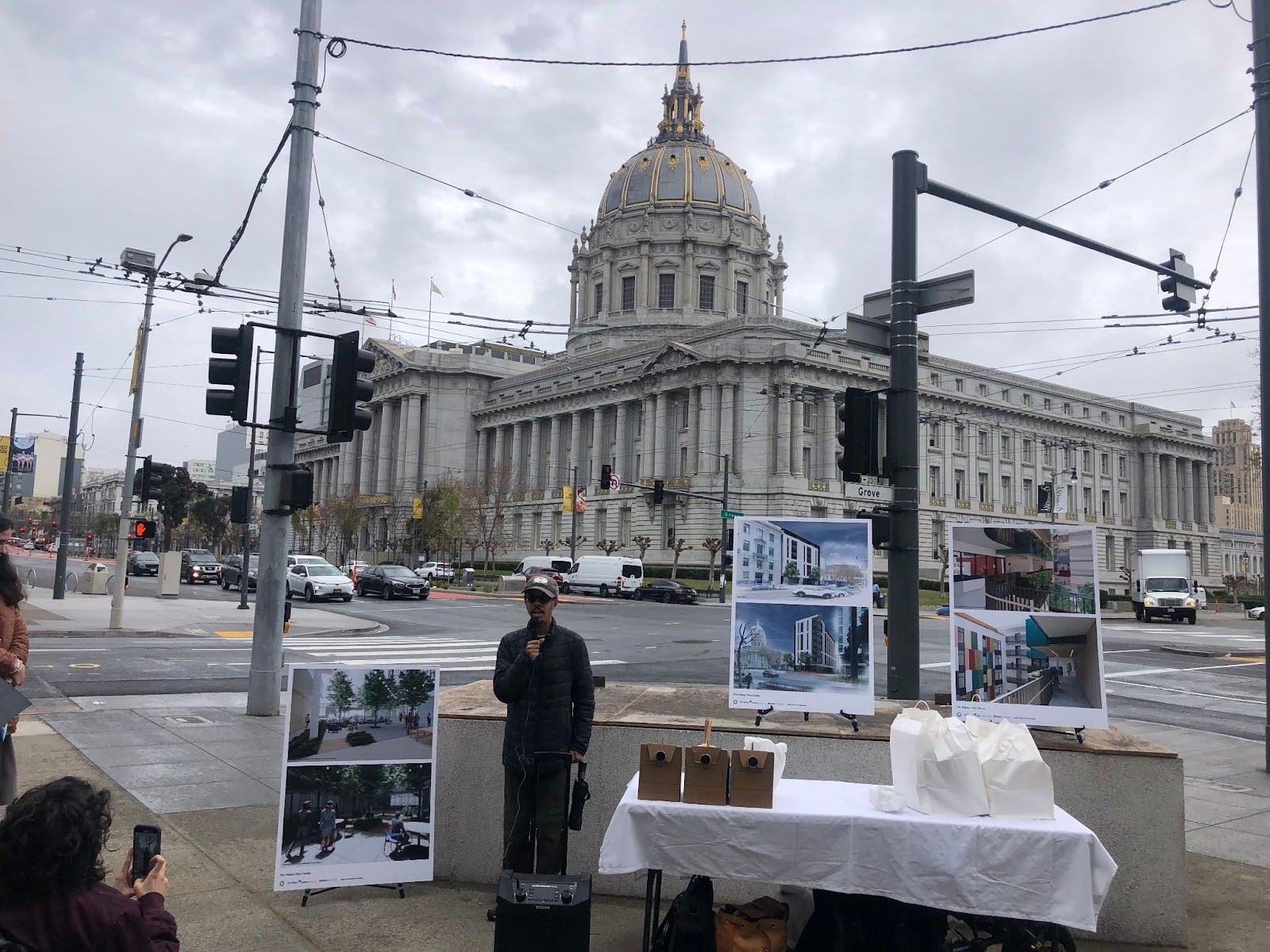 Isaac Haney Owens speaks in front of San Francisco City Hall. There is a table on one side of him and renderings of The Kelsey Civic Center behind him