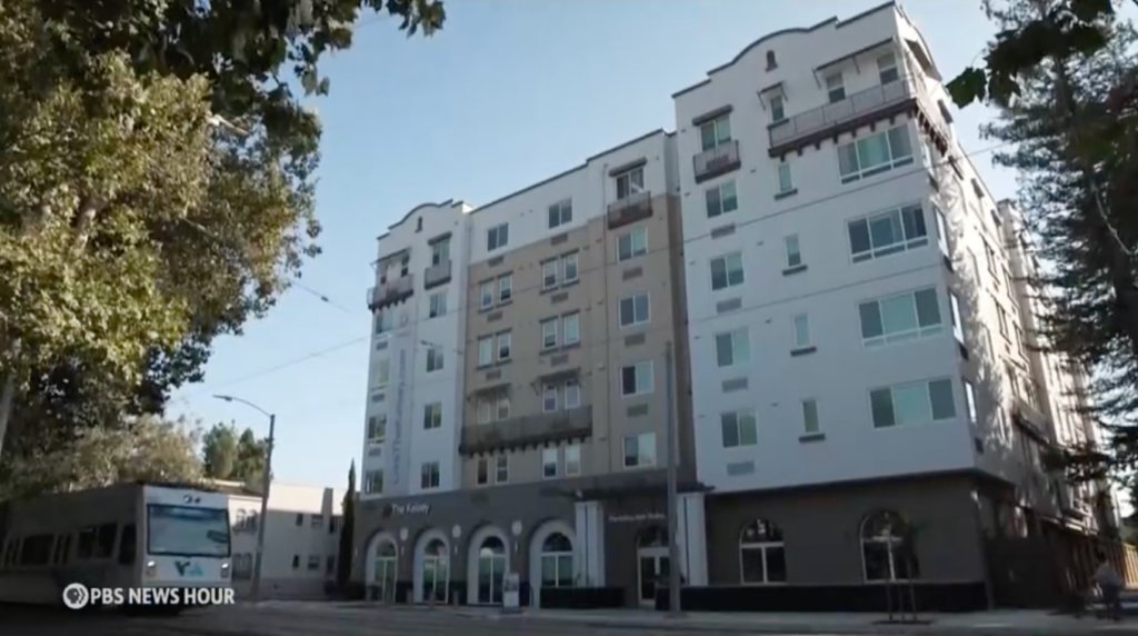 A screenshot of the exterior of The Kelsey Ayer Station with a light rail train passing it. The PBS News Hour logo is in the lower left corner.