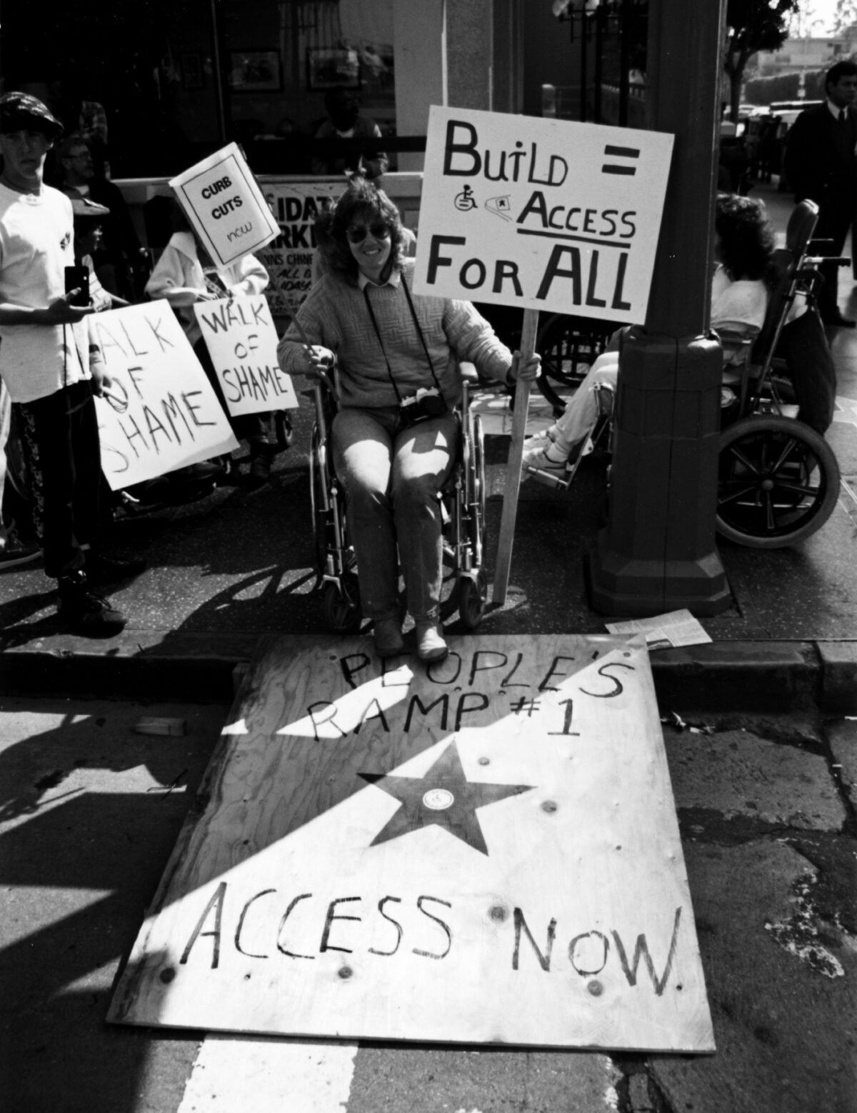 A black-and-white photo of people protesting. Some are in wheelchairs and some are standing. One person is in a wheelchair and holding a sign that says, “Build = Access For All,” and another sign that says, “Curb Cuts Now.” They are using a ramp made out of a piece of wood, that reads, “People’s Ramp #1. Access Now.” The ramp has a star with the international symbol of access in the middle of it.